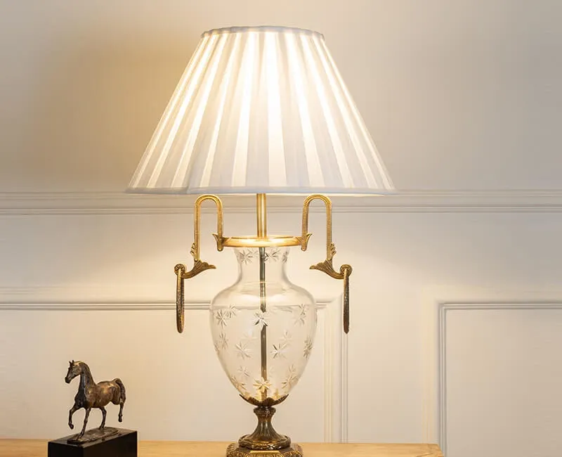 Luxury decorative table lamp featuring an engraved transparent glass vase base, antique gold metal accents and an elegant white pleated fabric lampsha
